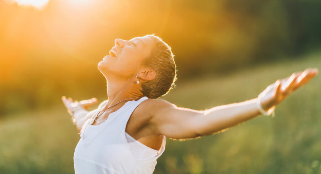 Close up of someone in a field, with short hair, in a white vest, head inclined upwards to the sun, smiling, arms outstretched horizontally to the side, palms up